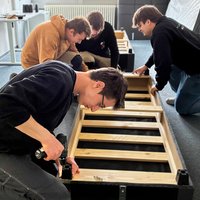 Auszubildende der Firma Mosca bauen mit Auszubildenden des Berufsbildungswerks Mosbach-Heidelberg in einem Zimmer Möbel zusammen.